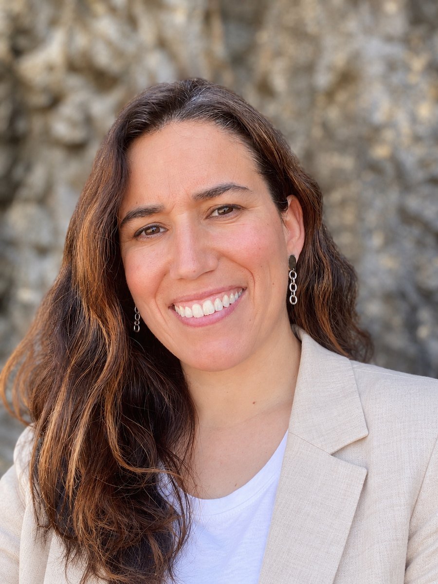 Natalia L. Salas sonriendo, con el pelo largo castaño y chaqueta clara, frente a un muro de piedra